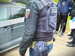 Na zdjęciu widać fragment sylwetki policjanta, z lewej fragment samochodu a dalej innych policjantów prowadzących zatrzymanego mężczyznę.