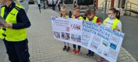 na zdjęciu dzieci trzymają dwa plakaty informujące o zmianach w przepisach ruchu drogowego. Przy nich stoi policjantka.